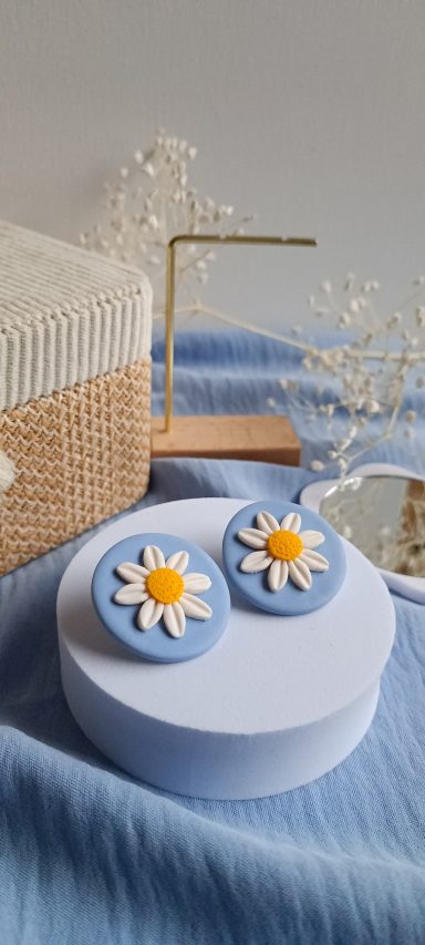 Boucles d'oreilles bleues en forme de fleurs avec un centre jaune, posées sur une surface douce.